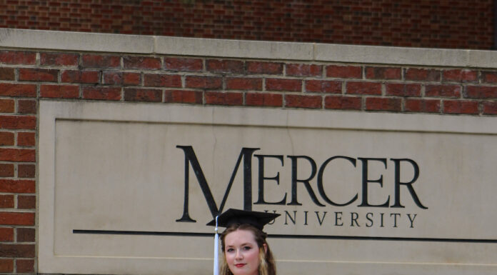 Graduate Spotlight: Ella Bissonnette Ella Bissonnette in cap and gown stands in front of a Mercer University sign and brick wall.
