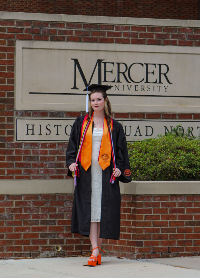 Ella Bissonnette in cap and gown stands in front of a Mercer University sign and brick wall.