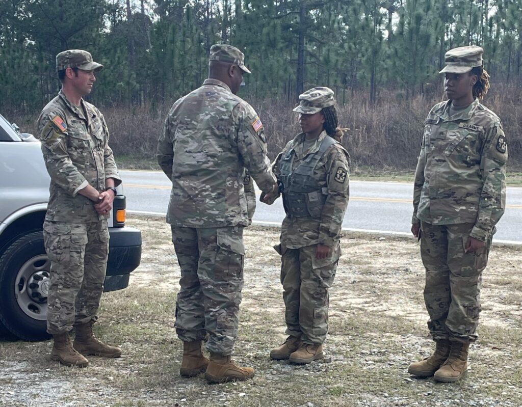 Four military personnel in uniform stand outside near a road; two are shaking hands.