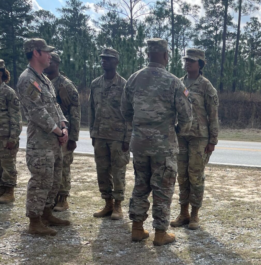 Group of soldiers in uniform standing on a dirt path near a road, with trees and clouds in the background.
