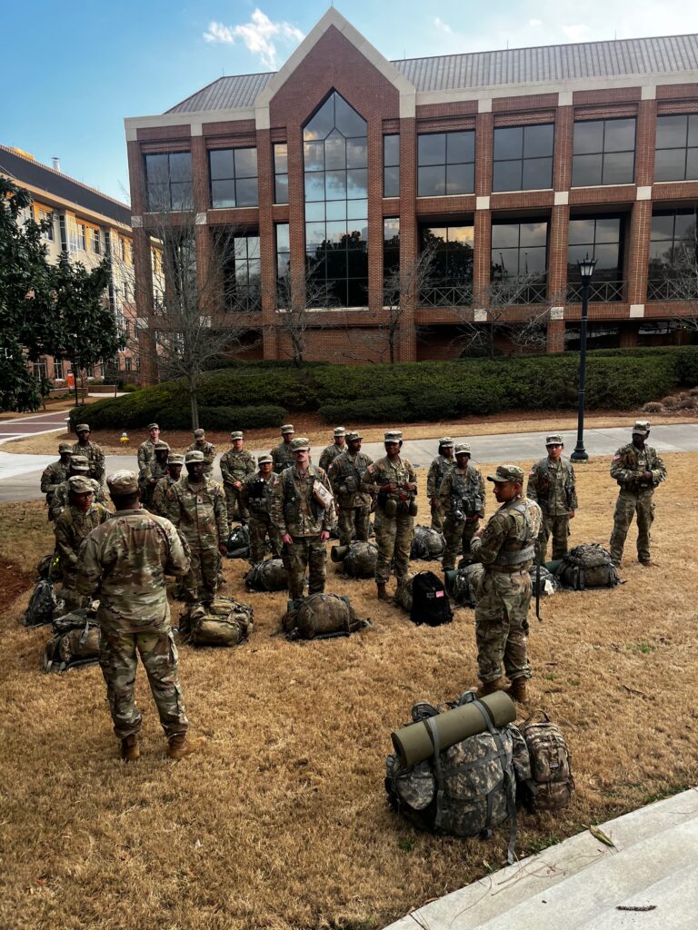 A group of uniformed military personnel stand in a circle on a grassy area with backpacks and gear, near a multi-story building.