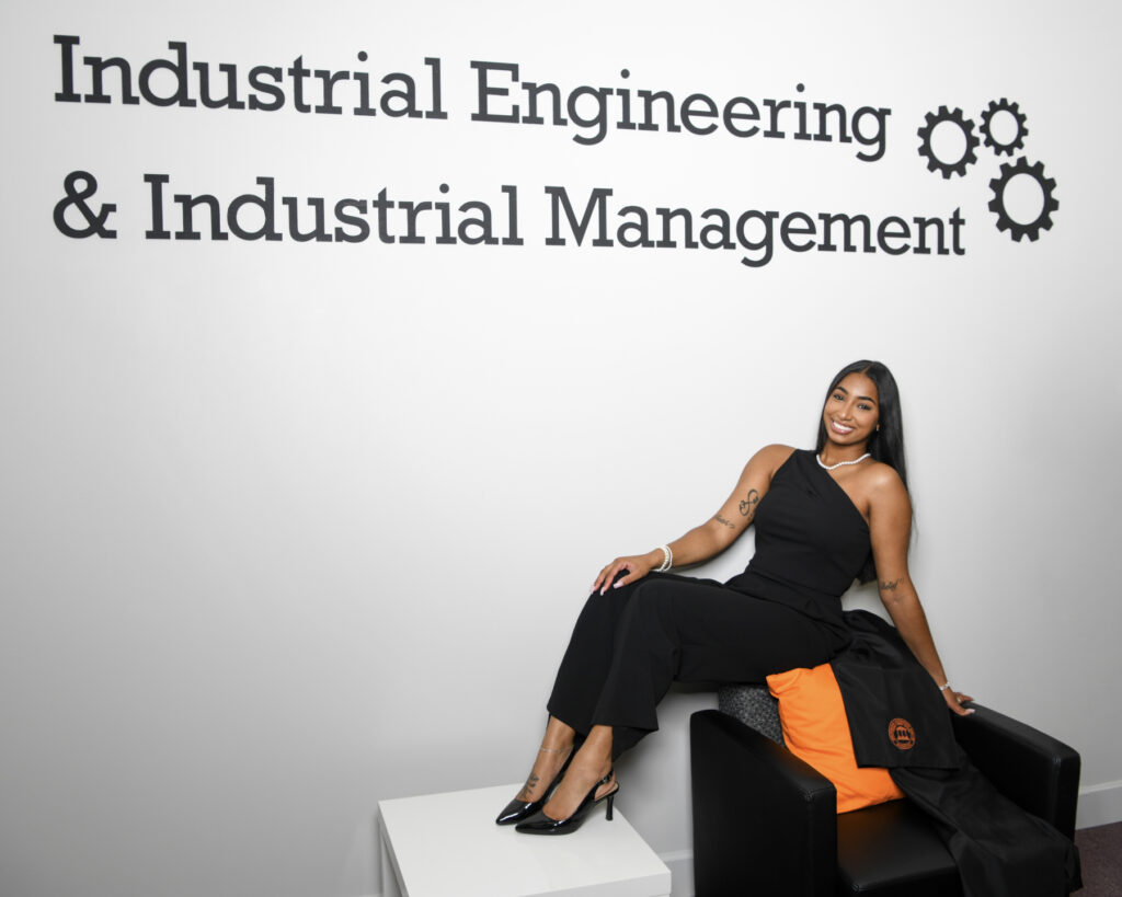 Imani Choukair sitting on a chair in front of a wall with the text: Industrial Engineering & Industrial Management.