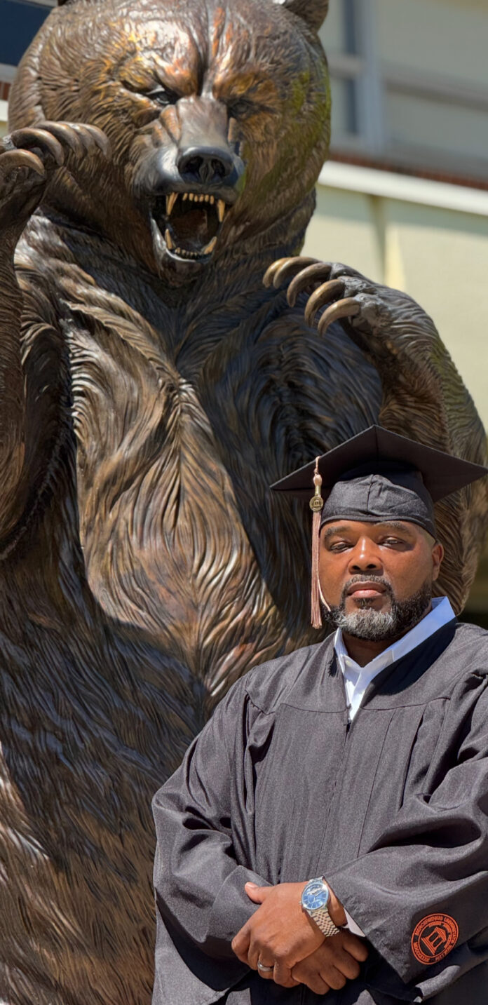 James Stephens, em um vestido de formatura e boné, fica na frente de uma grande estátua de urso.