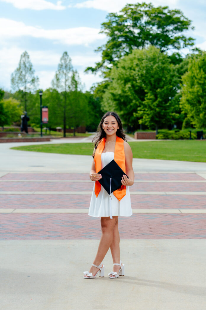 Jonalyn Hidalgo, in a white dress and orange graduation stole, holds a cap while standing outdoors on a paved walkway.