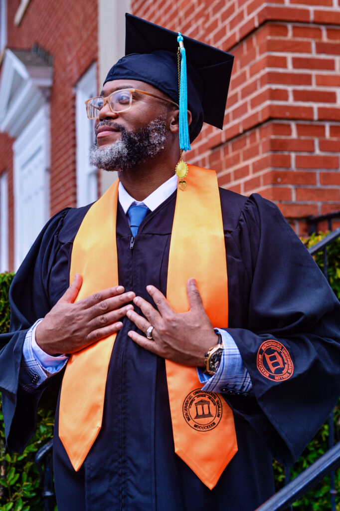 Ladji Ruffin, in graduation attire, stands in front of a brick building, wearing an orange stole and a mortarboard.