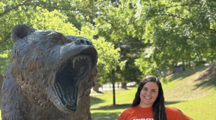 Graduate Spotlight: Lennox Bell Lennox Bell wears a Mercer Bears shirt and stands beside a large bear statue outdoors on a sunny day.