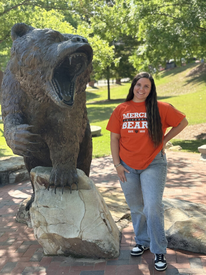 Lennox Bell wears a Mercer Bears shirt and stands beside a large bear statue outdoors on a sunny day.