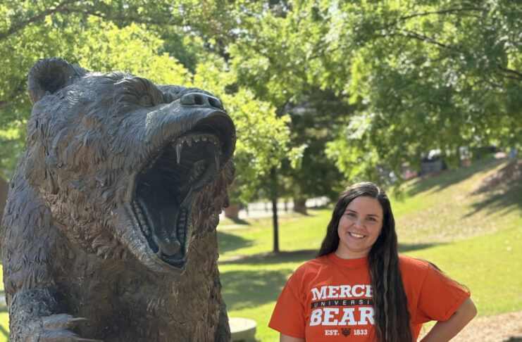 Lennox Bell wears a Mercer Bears shirt and stands beside a large bear statue outdoors on a sunny day.