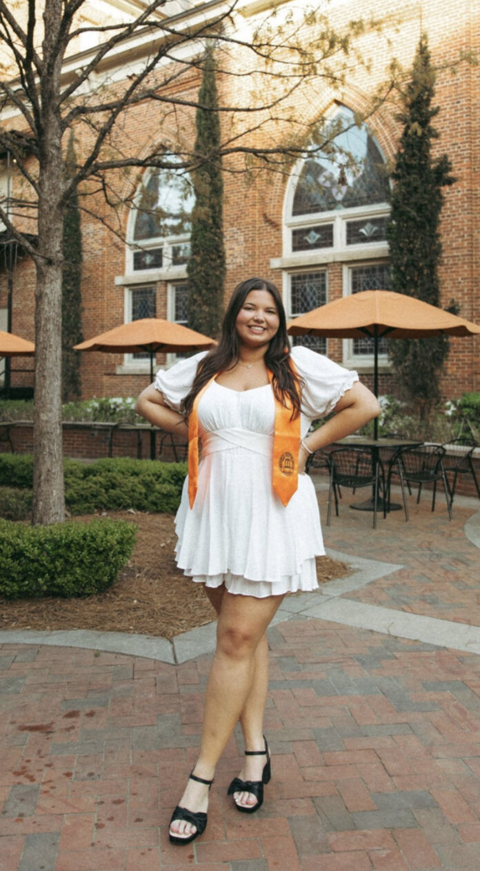 Lucy Roach in a white dress and orange graduation stole stands and smiles in a courtyard with tables and umbrellas.