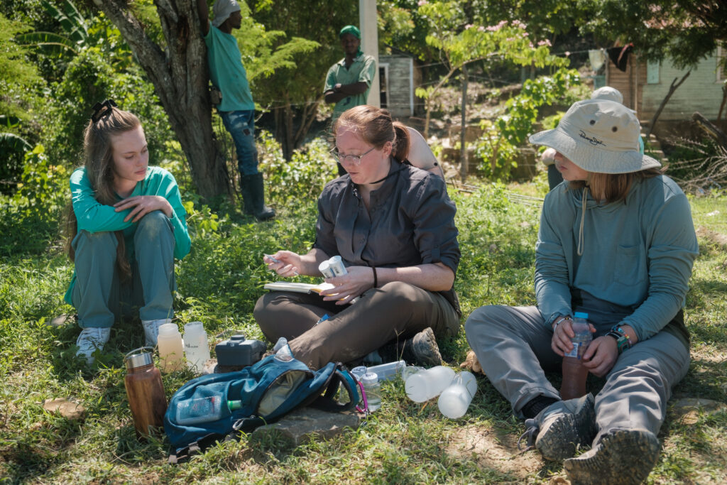 Three people sit on grass outdoors analyzing water samples with scientific equipment and bottles placed in front of them.