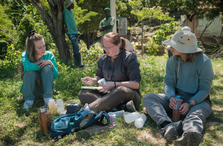 Three people sit on grass outdoors analyzing water samples with scientific equipment and bottles placed in front of them.