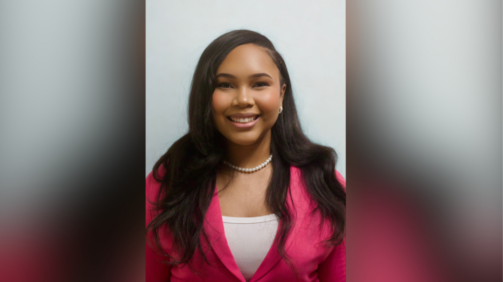 A woman with long dark hair and a pink blazer smiles at the camera in a portrait with a plain background.