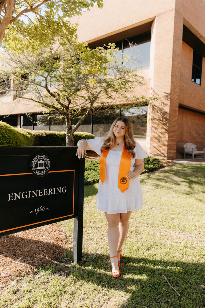 Rose Gambrell in a white dress and orange stole stands by Mercer's engineering building sign.