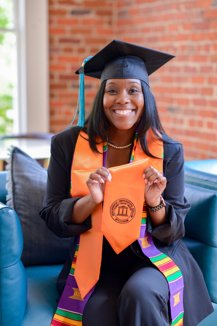 Tamia Sammons, in graduation attire, smiles while seated, holding an orange stole with a Mercer emblem.