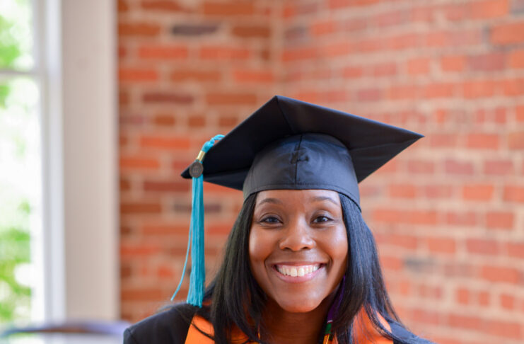 Tamia Sammons, in graduation attire, smiles while seated, holding an orange stole with a Mercer emblem.