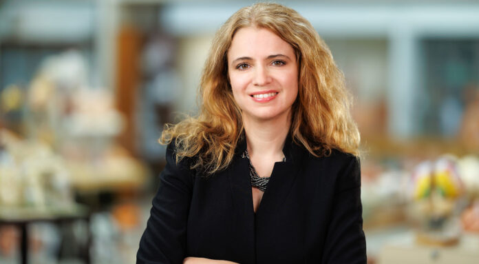Smiling woman with long curly blonde hair and a black blazer stands in the School of Medicine library with a blurred background.