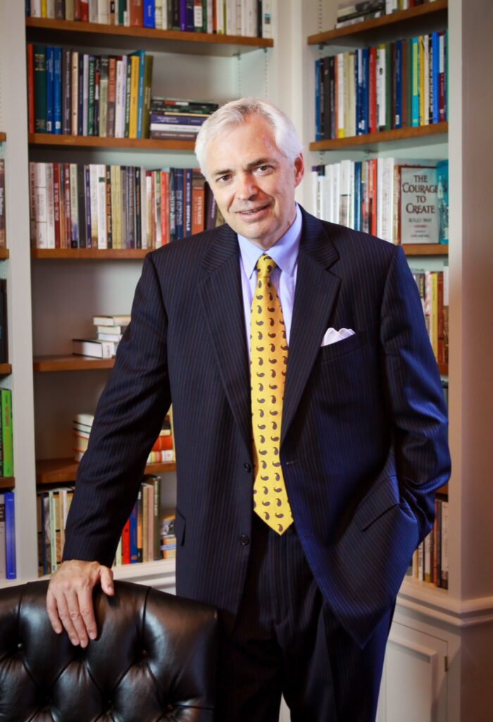 A man in a suit with a yellow tie stands in a library, resting one hand on a leather chair.