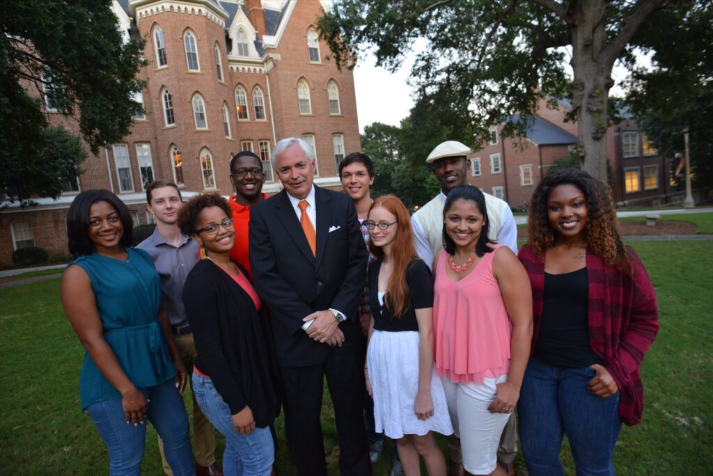 A group of people pose for a photo outside in front of a large brick building with trees and grass visible.