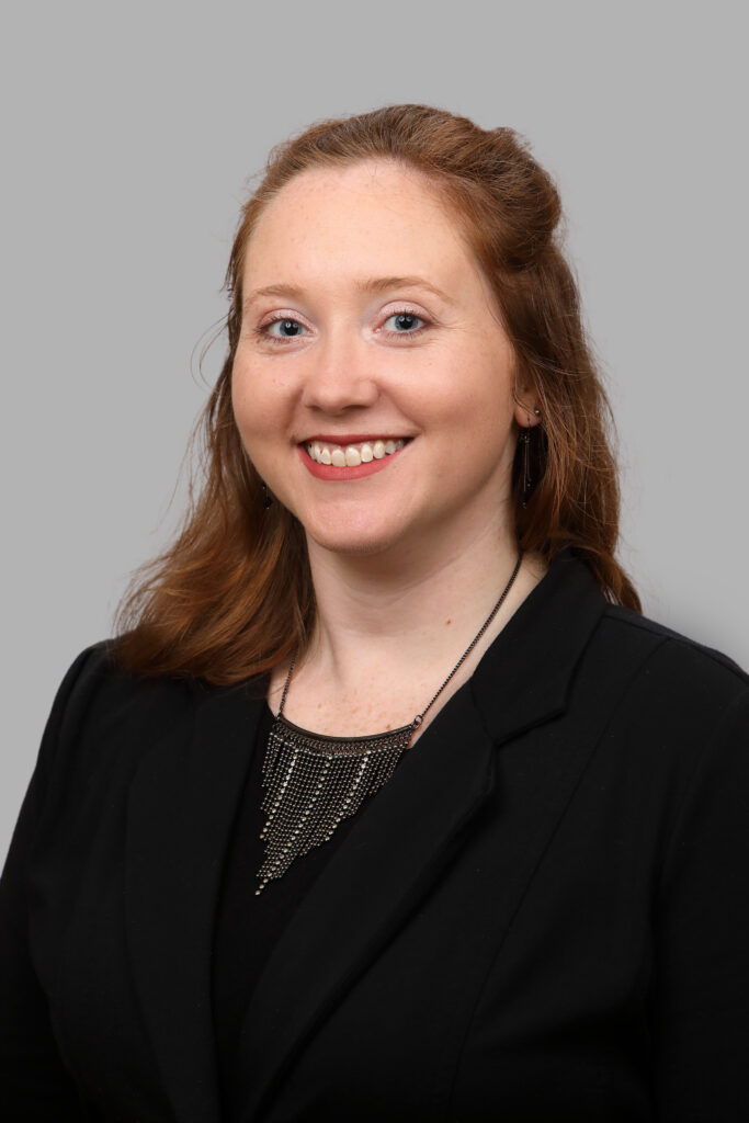 Woman with long red hair smiling, wearing a black blazer and necklace, posed against a plain gray background.