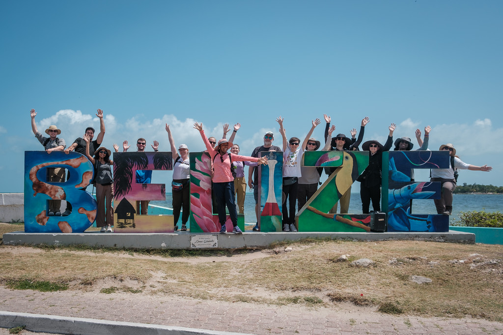A group of people standing and raising their arms in front of large, colorful “BELIZE” letters by the water.