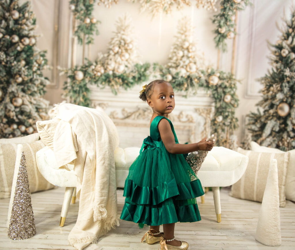 A young girl in a green dress stands by a white bench in a festive, holiday-themed room with winter decorations.