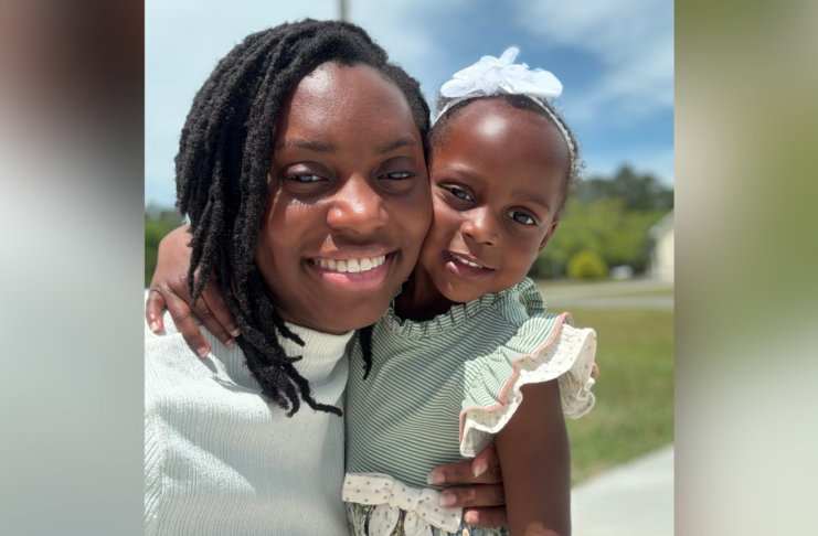 An adult and a young child smile outdoors, with the child wearing a white bow and green striped dress.