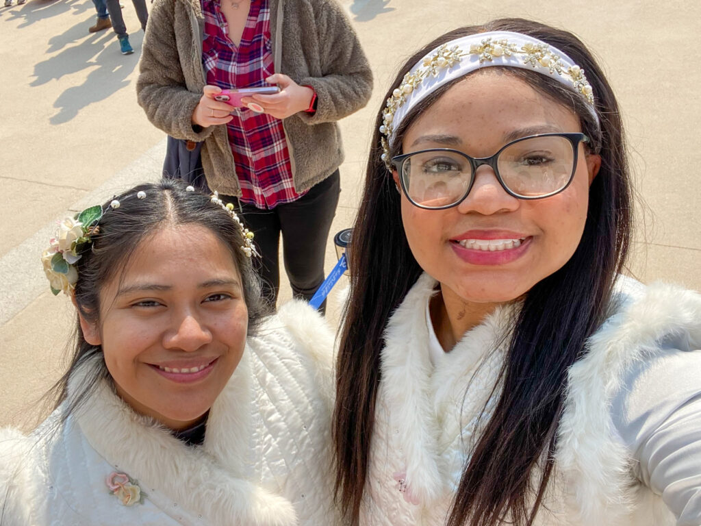 Two women smiling, wearing floral headbands and white fur-trimmed garments.