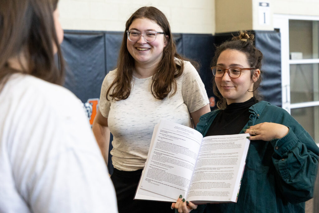 Two people stand and smile while showing an open book to a third person in an indoor setting.