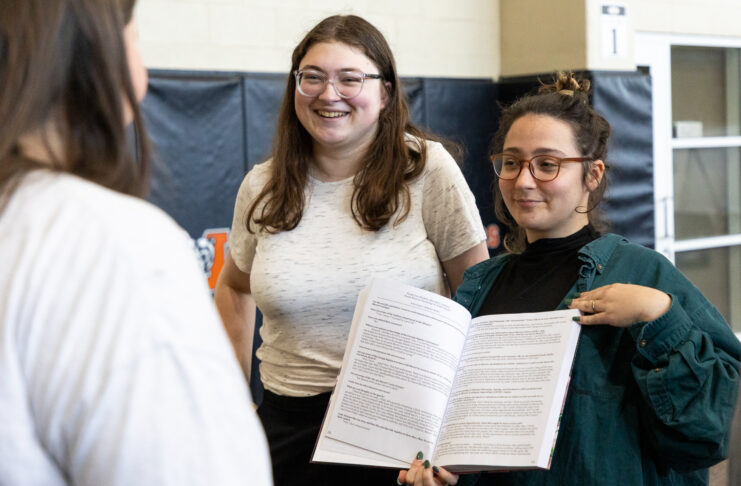 Two people stand and smile while showing an open book to a third person in an indoor setting.