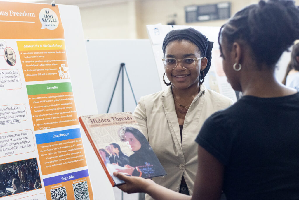 Two women stand by a presentation board; one hands the other a book titled Hidden Threads.