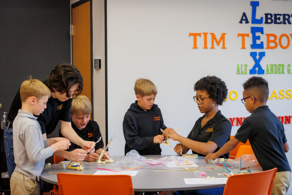 Six children work together on a craft project at a round table in a classroom with colorful letters on the wall behind them.
