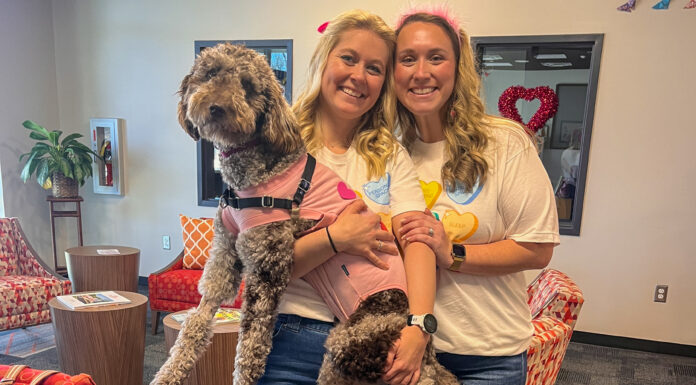 A paw-some surprise: How an abandoned pup became Roberts Academy’s newest superhero Caroline Grove and Elizabeth Eidson stand indoors smiling and holding a large curly-haired dog wearing a pink harness.