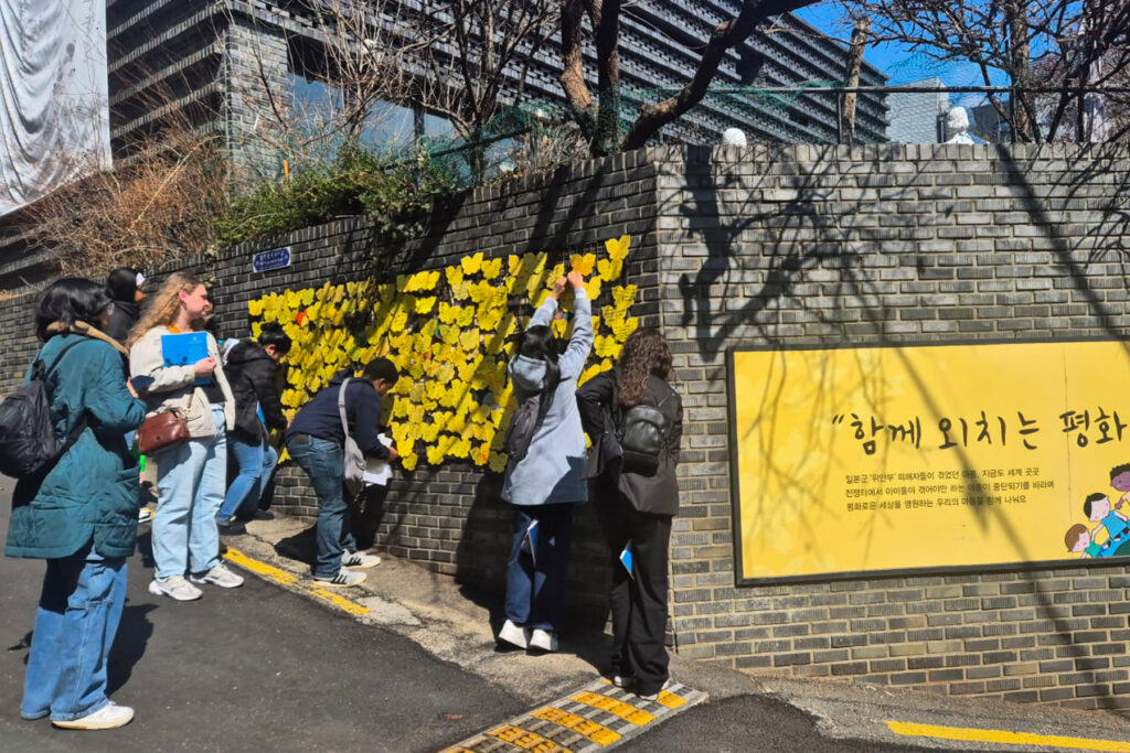People placing yellow paper butterflies on a brick wall beside a large yellow sign on a sunny day.