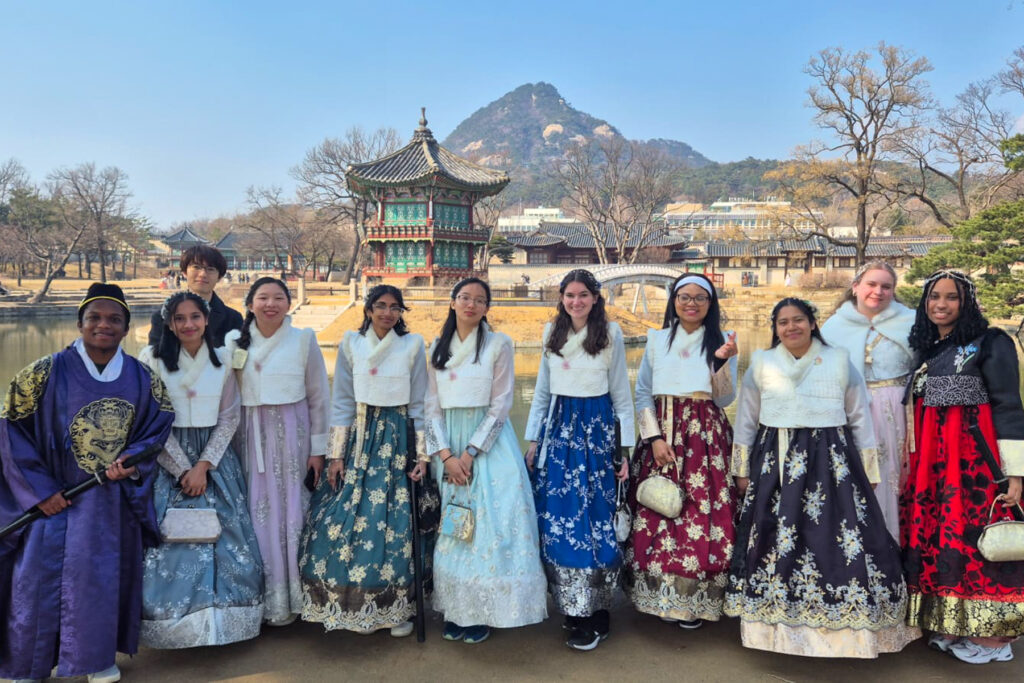 Group of people in traditional Korean hanbok posing in front of a pavilion with a mountain backdrop on a clear day.