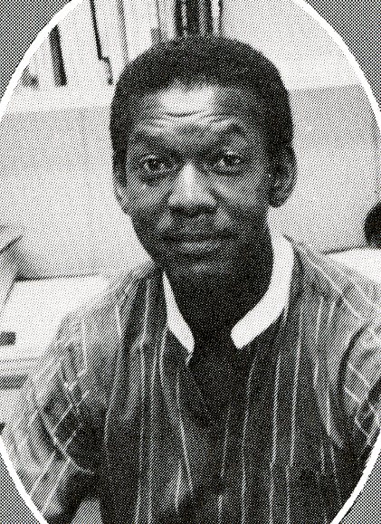A man in a striped shirt sits at a desk, looking at the camera; books and papers are visible in the background.