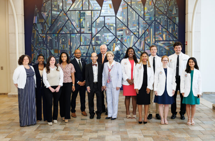 A group of people, some in white coats, pose for a photo indoors in front of a large, colorful stained glass mural.