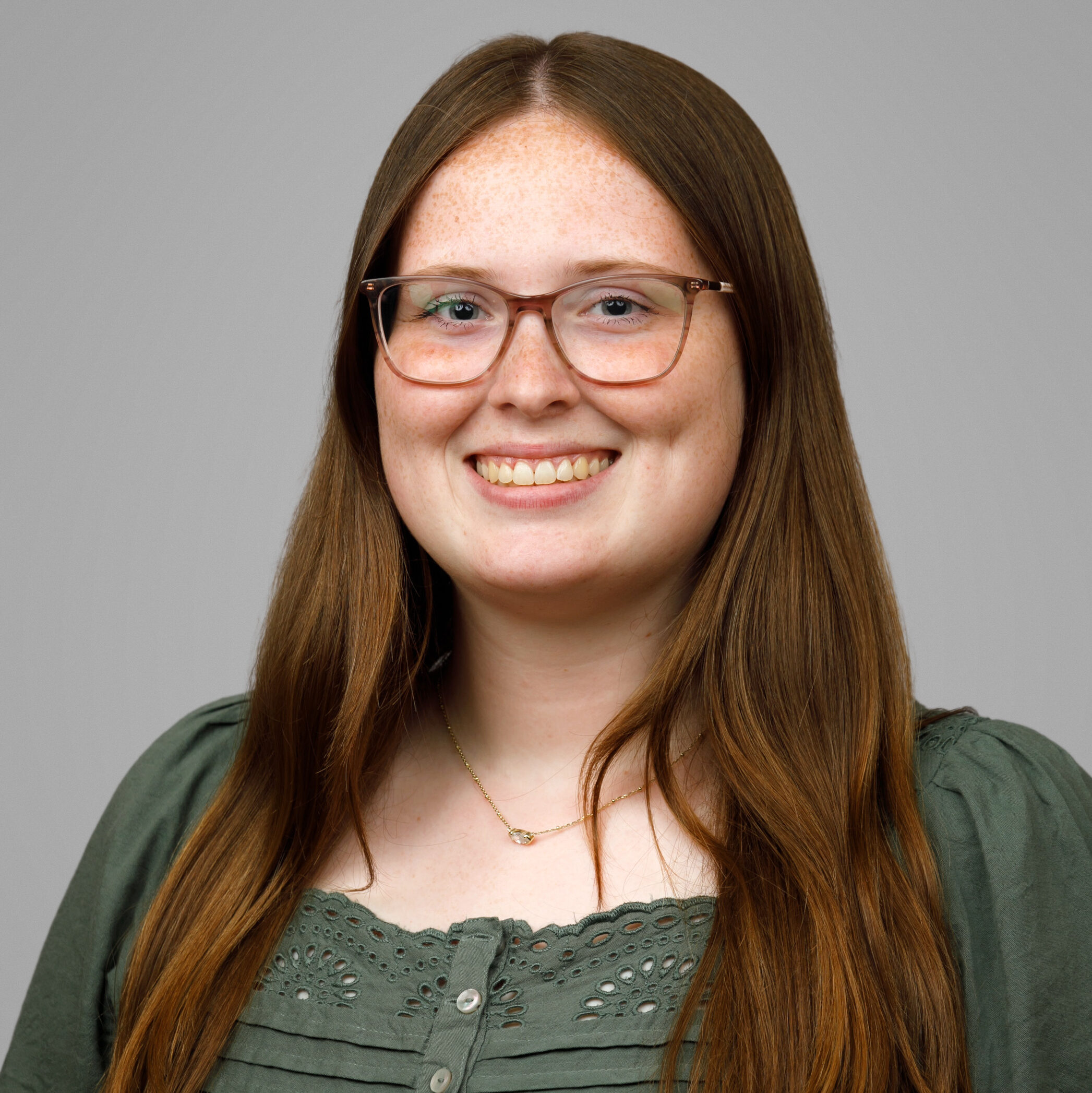 A young woman with long brown hair, glasses, and a green blouse smiles in front of a plain gray background.