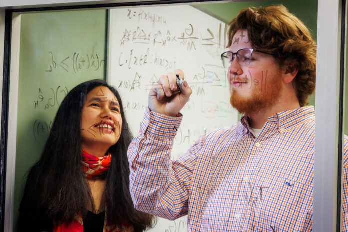250409MUGoldwaterKinney_0009 Dr. Makhin Thitsa and Wesley Kinney work at a glass window covered with mathematical equations. Kinney is writing while Dr. Thitsa observes.