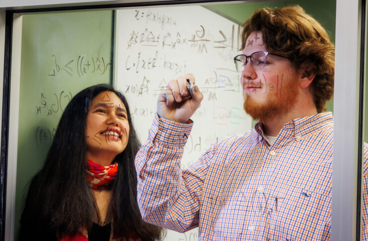 Dr. Makhin Thitsa and Wesley Kinney work at a glass window covered with mathematical equations. Kinney is writing while Dr. Thitsa observes.