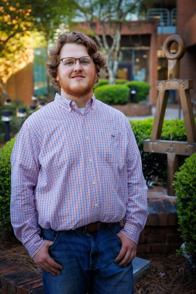 Wesley Kinney stands outdoors near a metal sculpture and green shrubs.