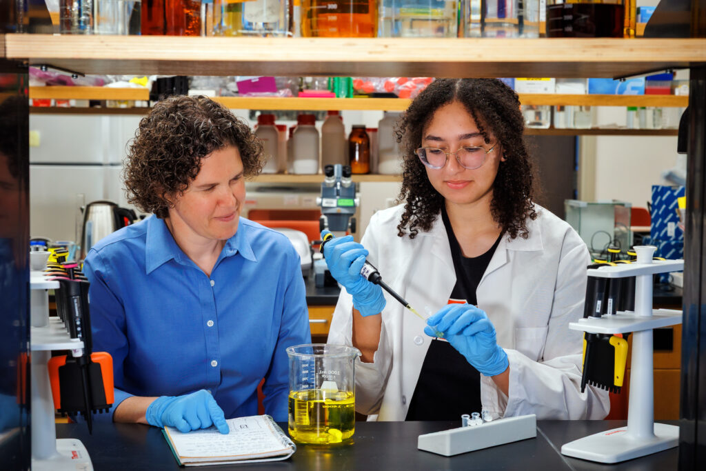 Two people in a laboratory, one taking notes while the other uses a pipette to handle liquid in a beaker.