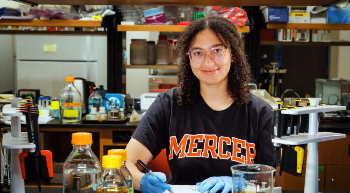 Goldwater Scholar is passionate about neuroscience, research A person wearing gloves and a Mercer shirt writes notes at a lab table with bottles, beakers, and pipettes.