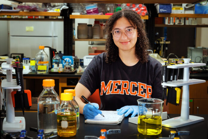 A person wearing gloves and a “Mercer” shirt writes notes at a lab table with bottles, beakers, and pipettes.
