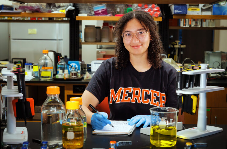 A person wearing gloves and a “Mercer” shirt writes notes at a lab table with bottles, beakers, and pipettes.