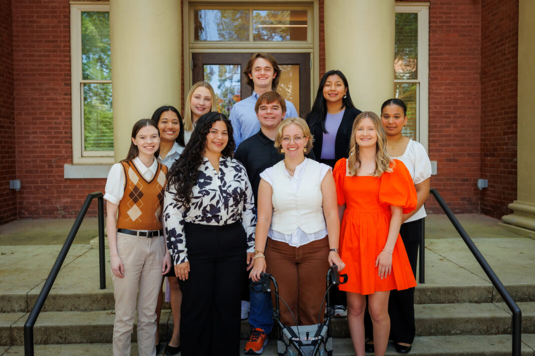 Ten people pose and smile for a group photo on outdoor steps in front of a brick building with columns.