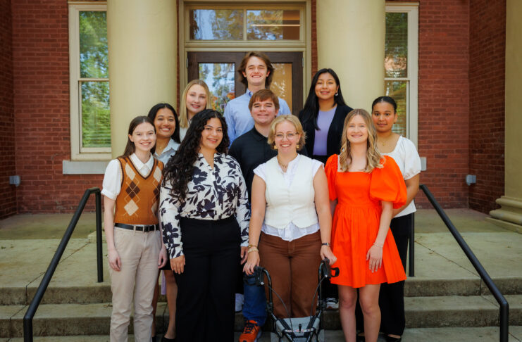 Ten people pose and smile for a group photo on outdoor steps in front of a brick building with columns.