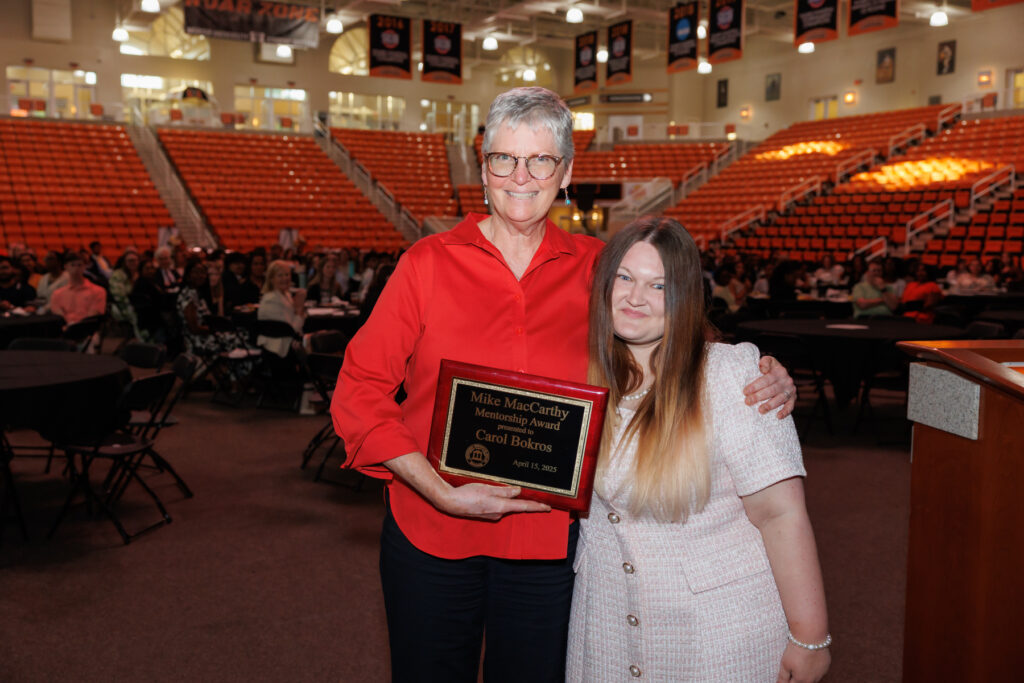 Carol Bokros and Johna Wright stand together in an auditorium. Carol Bokros holds a plaque that reads Mike MacCarthy Membership Award.