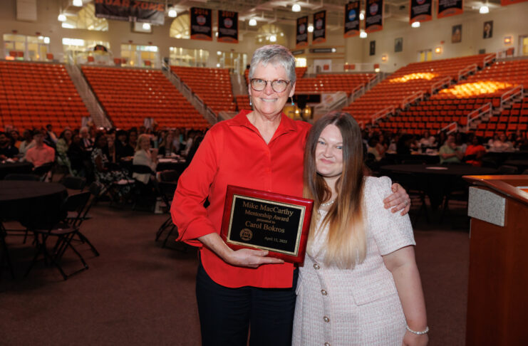Carol Bokros and Johna Wright stand together in an auditorium. Carol Bokros holds a plaque that reads Mike MacCarthy Membership Award.