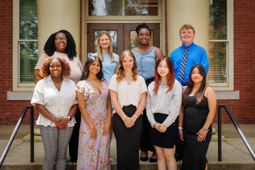 Nine people pose for a group photo on steps outside a brick building with white columns and glass doors.