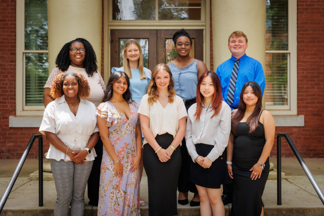 Nine people pose for a group photo on steps outside a brick building with white columns and glass doors.